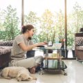 A woman sits on the floor in front of a low table using a phone and laptop computer, a dog is next to her.